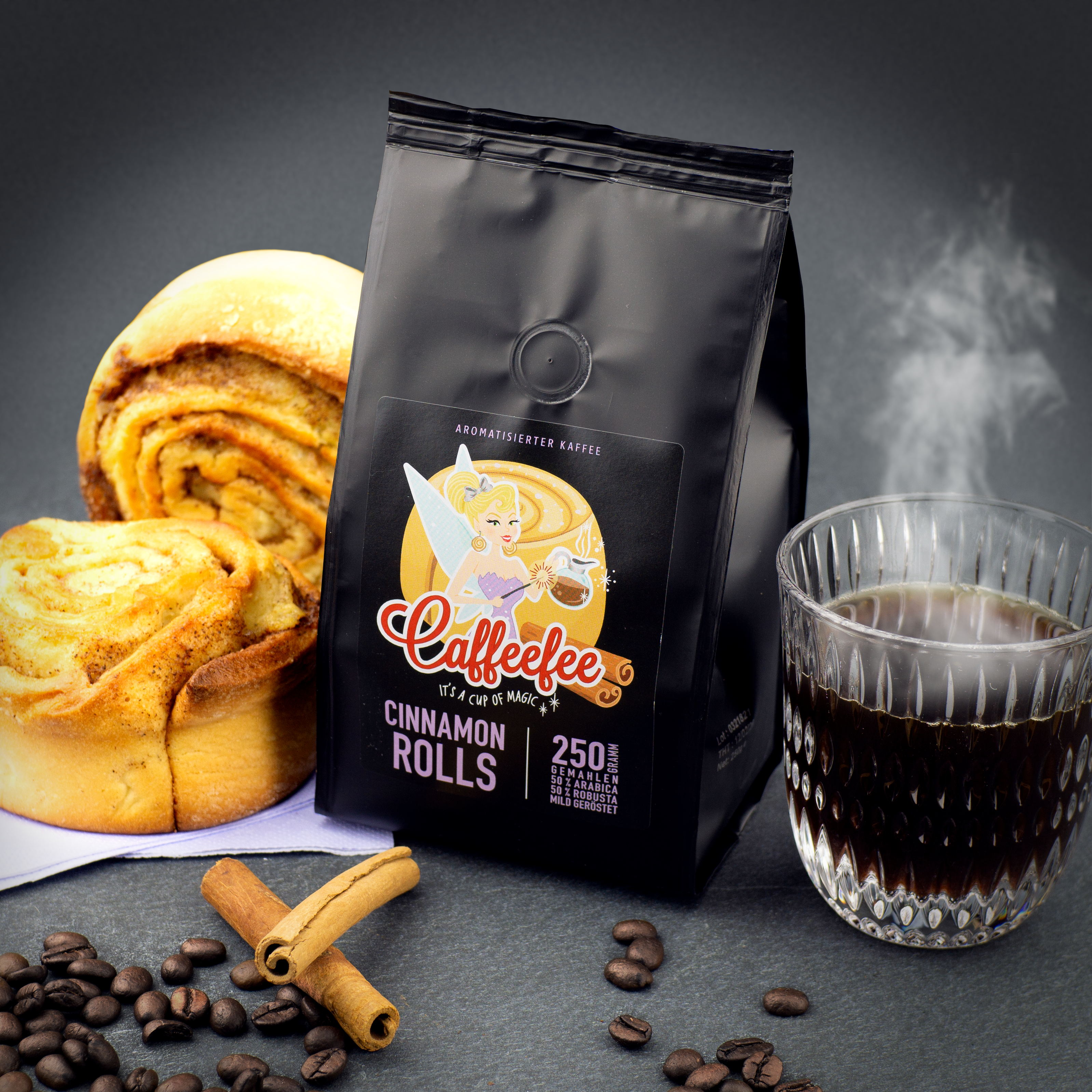 Päckchen Caffeefee Cinnamon Rolls, daneben zwei Zimtschnecken und eine Tasse mit frisch gebrühten Kaffee. Im Vordergrund Kaffeebohnen und Zimtstangen.