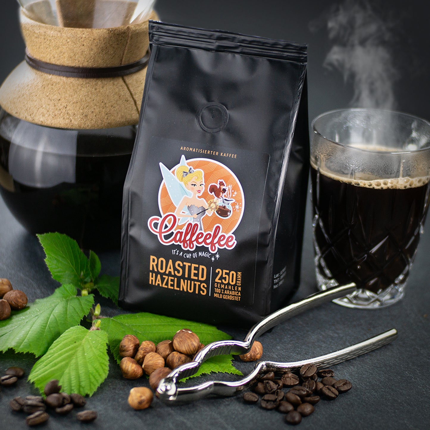 Abbildung Packung Caffeefee Roasted Hazelnuts. Rechts daneben eine Tasse mit heißem Kaffee. Links im Hintergrund eine gläserne Kaffeekanne mit Korkgriff. Vorne liegen Haselnüsse, Haselnussblätter, ein silberner Nussknacker und Kaffeebohnen.